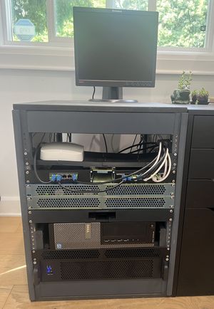 A homemade server rack holding several servers, networking equipment, and an old monitor on top. The rack sits beside office desk storage, and in front of a window with visible trees outside